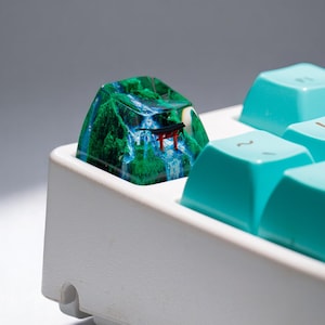 Puede incluir: Una tecla de teclado personalizada que presenta un paisaje en miniatura con una cascada, &aacute;rboles verdes y una peque&ntilde;a puerta torii roja. La tecla est&aacute; colocada en un teclado blanco con teclas turquesas.