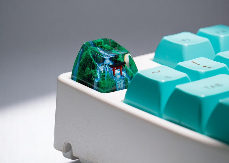 Puede incluir: Una tecla de teclado personalizada que presenta un paisaje en miniatura con una cascada, &aacute;rboles verdes y una peque&ntilde;a puerta torii roja. La tecla est&aacute; colocada en un teclado blanco con teclas turquesas.