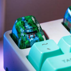 Puede incluir: Primer plano de teclas de teclado personalizadas con un dise&ntilde;o esc&eacute;nico. Las teclas presentan una resina transparente con un paisaje detallado de una cascada, &aacute;rboles y una puerta torii roja. Las teclas circundantes son de color verde azulado claro.