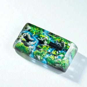 Puede incluir: Objeto rectangular de resina transparente con un dise&ntilde;o de paisaje. El dise&ntilde;o presenta elementos verdes, azules y blancos que se asemejan a un r&iacute;o y follaje circundante. Un objeto rectangular negro est&aacute; incrustado en la resina.