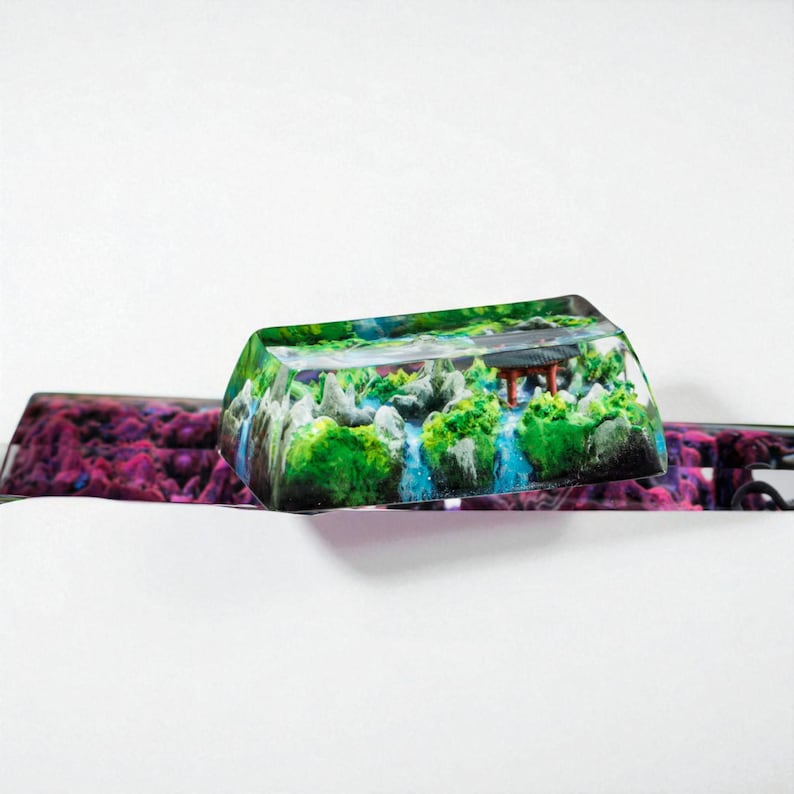 Puede incluir: Una tecla rectangular de resina transparente con una escena de paisaje en miniatura. La escena incluye &aacute;rboles verdes, cascadas y una peque&ntilde;a estructura con techo rojo. La tecla est&aacute; encima de un teclado con un dise&ntilde;o p&uacute;rpura y rosa.