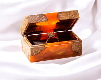 Op de afbeelding: Een houten ringendoos met een transparant oranjehars deksel. De doos is open en er ligt een diamanten ring in.