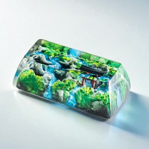 Puede incluir: Una tecla rectangular de resina transparente con un paisaje en miniatura. La escena incluye una cascada, vegetaci&oacute;n verde y una puerta torii roja y negra. Un art&iacute;culo decorativo &uacute;nico.
