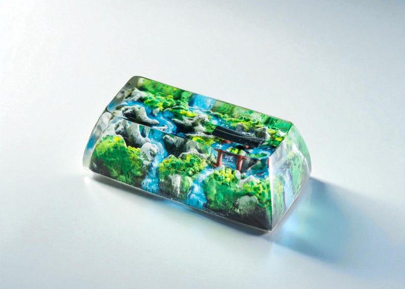 Puede incluir: Una tecla rectangular de resina transparente con un paisaje en miniatura. La escena incluye una cascada, vegetaci&oacute;n verde y una puerta torii roja y negra. Un art&iacute;culo decorativo &uacute;nico.