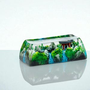 Puede incluir: Una tecla rectangular de resina transparente con una escena de paisaje en miniatura. La escena incluye una pagoda roja, follaje verde, cascadas y rocas grises. La tecla est&aacute; dise&ntilde;ada para un teclado mec&aacute;nico.
