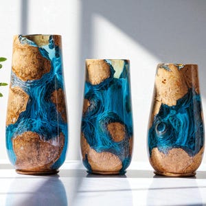Puede incluir: Tres jarrones decorativos con un diseño único. Los jarrones presentan una combinación de madera natural y resina azul vibrante, creando un contraste llamativo. La madera es de color marrón claro, mientras que la resina tiene una apariencia arremolinada, similar al océano. Los jarrones son de diferentes alturas.