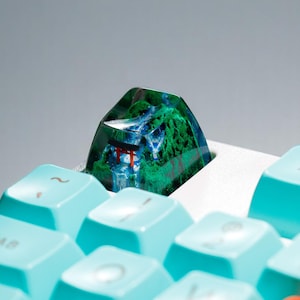 Puede incluir: Una tecla de teclado personalizada con un paisaje en miniatura. Presenta una puerta torii roja, una cascada y follaje verde. La tecla se encuentra sobre un fondo de teclas de teclado azul claro.