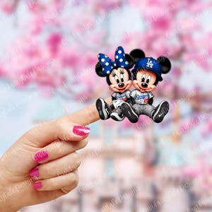Puede incluir: Un par de personajes de dibujos animados de Mickey Mouse y Minnie Mouse que llevan uniformes de béisbol de los Dodgers de Los Angeles. Minnie Mouse lleva un uniforme de béisbol azul y blanco con un lazo rojo y lunares blancos. Mickey Mouse lleva un uniforme de béisbol azul y blanco con una gorra de béisbol azul que dice "LA". Los personajes están sentados en un dedo.