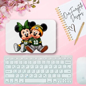Può includere: Un tappetino per mouse bianco con un'immagine di cartone animato di Topolino e Minnie Mouse che indossano maglie da calcio verdi e dorate. Le maglie hanno il numero 10.