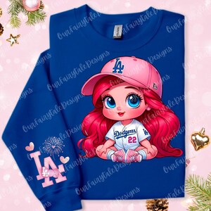 May include: A blue long-sleeved shirt with a pink and white cartoon image of a girl wearing a baseball cap with the letters "LA" and a baseball jersey with the text "Dodgers 22". The shirt has a pink heart and fireworks design on the sleeve.