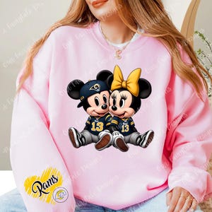 Op de afbeelding: Roze sweatshirt met een afbeelding van Mickey en Minnie Mouse in American football-shirts met nummer 13. Mickey draagt een marineblauwe pet. De sweatshirt heeft een Rams-logo op de mouw.