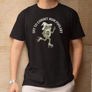 May include: Black t-shirt featuring a cartoon frog on roller skates, wearing a hat and holding a coffee cup, with the text "OFF TO COMMIT MOM FOOLERY" in a semi-circular design. The frog is making a peace sign.