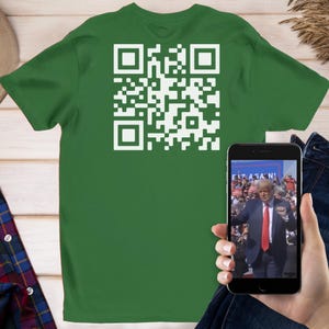 May include: A green t-shirt with a white QR code on the back. A person is holding a phone showing a video of a man in a suit with the text "Trump Again!" on the screen.