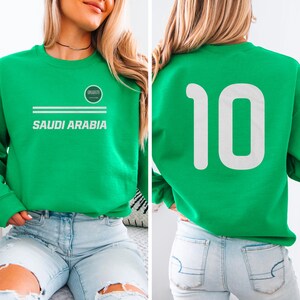 A woman wearing a green sweatshirt with the number 10 on it, and a logo that says "SAUDI ARABIA" printed on it.
