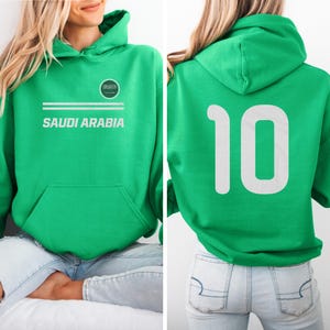 A woman wearing a green hoodie with the text "SAUDI ARABIA" and the number "10" on the back.