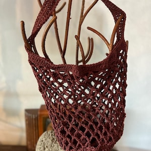 May include: A burgundy crocheted net bag with a shoulder strap. The bag is suspended from a decorative branch structure. The open weave design is visible, and the bag appears to be empty. The bag is a deep reddish-brown color.