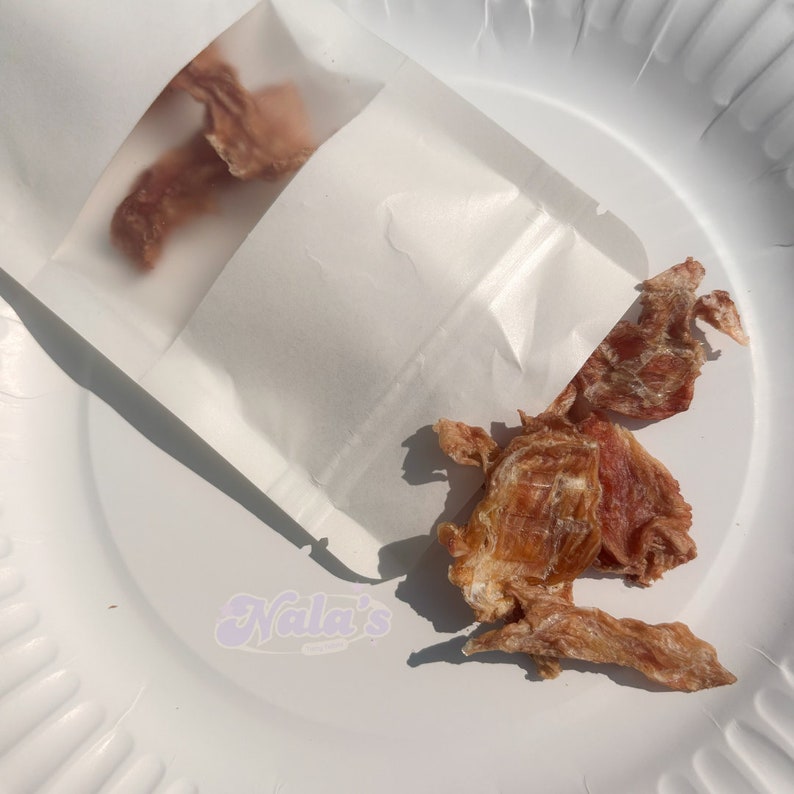 Pu&ograve; includere: Un sacchetto di carta bianco pieno di bocconcini di carne secca per cani. Il sacchetto &egrave; aperto e i bocconcini cadono su un piatto bianco.