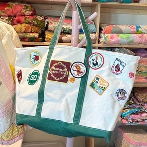 Puede incluir: Un bolso tote de lona blanca con bordes y asas de color verde oscuro, decorado con varios parches bordados coloridos que incluyen los logotipos de Hawaiian Queen Coffee, Makapuu Point, Volcano House y Matsumoto Shave Ice en el frente.