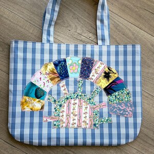 Peut inclure: Un sac fourre-tout à carreaux bleus et blancs avec un motif de patchwork coloré représentant un arc-en-ciel et une maison. Le motif est cousu sur le devant du sac.