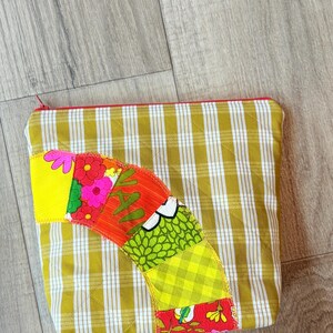 Peut inclure: Une pochette zippée à carreaux jaunes et blancs avec un motif patchwork coloré sur le devant. Le motif patchwork présente une variété de couleurs, notamment le rouge, l'orange, le jaune, le vert et le bleu.