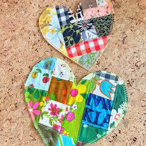 May include: Two handmade heart-shaped coasters. Each coaster is a patchwork of various fabric patterns, including floral, gingham, and geometric designs. The edges are stitched with contrasting thread. The coasters rest on a textured, light brown surface.