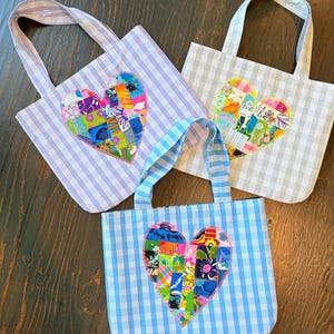 May include: Three tote bags made of gingham fabric in light purple, light gray, and light blue. Each bag features a central heart-shaped appliqué constructed from a colorful patchwork of various floral and geometric patterned fabric scraps.
