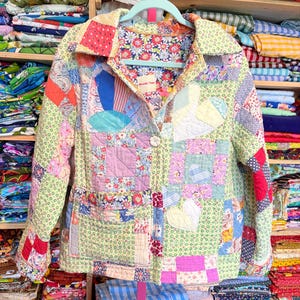 Puede incluir: Una chaqueta de manga larga confeccionada con varios parches de tela acolchada en colores pastel, incluyendo verde claro, rosa, amarillo y azul. La chaqueta presenta un cuello, un cierre de un solo botón y una etiqueta Prudy Nan en el interior.