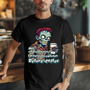 Pode incluir: Camiseta preta com um desenho animado de um zumbi segurando uma xícara de café. O zumbi tem pele verde, cabelo vermelho e uma jaqueta marrom. O texto na camiseta diz: "Not a morning person? I'm not even a person before coffee."