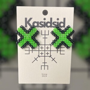 Op de afbeelding: Een paar groene en zwarte kralen oorbellen in de vorm van een X. De oorbellen worden getoond op een witte kaart met de tekst "Kasidsid" en "Since 2024".
