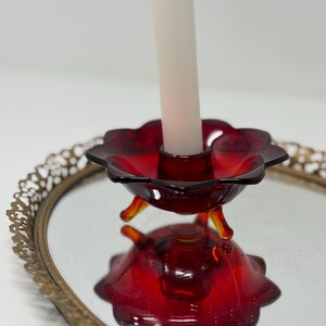 May include: A red glass candle holder with a flower-like design, holding a white candle. The candle holder sits on a mirrored surface, reflecting its shape. The holder has three small, yellow-orange feet. A gold-colored, ornate tray edge is visible.
