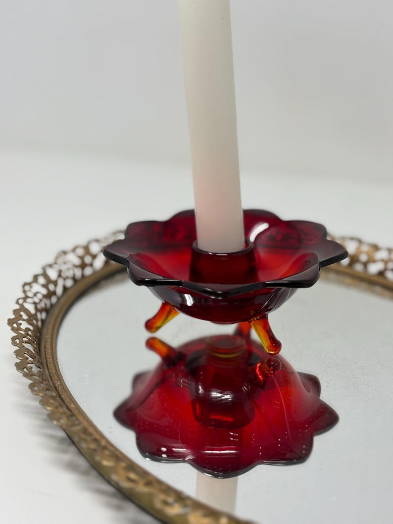 May include: A red glass candle holder with a flower-like design, holding a white candle. The candle holder sits on a mirrored surface, reflecting its shape. The holder has three small, yellow-orange feet. A gold-colored, ornate tray edge is visible.