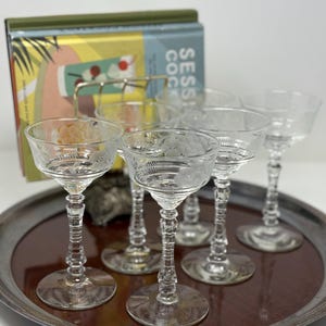 May include: A set of six clear, etched champagne glasses with detailed stems, arranged on a silver-rimmed tray. A cocktail book is in the background. The glasses have a delicate, vintage aesthetic.