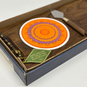 Può includere: Un vassoio di legno con un sottobicchiere arancione e viola, un coltello da formaggio e un utensile da portata con perline. Il sottobicchiere ha un design a mandala circolare. Un'etichetta verde recita "Hand Crafted of Selected Hardwoods."