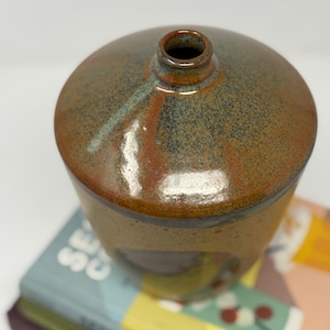 May include: A handmade ceramic vase with a rounded lid and a small opening. The vase has a speckled brown and tan glaze with hints of blue and red. The vase is placed on top of a stack of books.