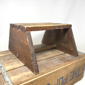 Op de afbeelding: Een kleine, donkerbruine houten opstapje met een rechthoekig bovenvlak en schuine zijsteunen. Het krukje heeft een verweerde afwerking met zichtbare houtnerf en gebruikssporen, en staat op een grotere houten kist met gedeeltelijke tekst.