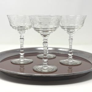 Könnte beinhalten: Ein Set aus vier klaren Glaskelchen mit geätztem Blumenmuster auf der Kuppa und einem mehrstufigen Stiel. Die Gläser sind auf einem runden, dunkel getönten Metalltablett mit einer glatten, reflektierenden Oberfläche angeordnet.