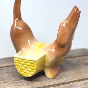 May include: A brown ceramic dachshund figurine with a glossy finish, featuring a yellow basket attached to its back. The dog is depicted in a standing position with its head tilted upward and tail pointed straight up, showcasing a smooth surface.