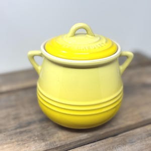 Puede incluir: Un tarro de gres amarillo brillante de Le Creuset con tapa a juego y dos asas laterales. El tarro presenta un cuerpo redondeado con tres anillos horizontales en relieve en la parte inferior y el nombre de la marca grabado en la tapa.