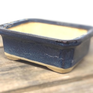 May include: A rectangular ceramic bonsai pot featuring a deep blue glazed exterior and a contrasting light beige interior. The vessel has a wide rim, slightly tapered sides, and four small feet at the base, providing a stable foundation.