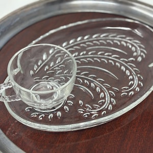 Könnte beinhalten: Ein Teetassen- und Untertassen-Set aus klarem Glas mit einem weizenähnlichen Design, das auf einer dunkelbraunen Oberfläche ruht. Das Set befindet sich auf einem silberumrandeten Tablett und erzeugt eine Vintage-Ästhetik. Die Tasse hat einen Henkel.