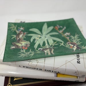 May include: A rectangular green glass tray with a jungle scene featuring monkeys and foliage, resting on a stack of books. The books have white and dark covers with gold lettering.