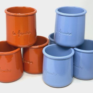 May include: A collection of six ceramic pots in orange and light blue, arranged in two groups. Each pot features a smooth, glossy finish and the embossed text la fermière on the front. The cylindrical vessels have a rounded rim and base.