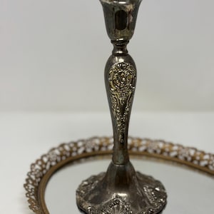 May include: A silver candlestick with ornate floral detailing. The candlestick has a flared top and a detailed base, sitting on a mirrored surface. The design features intricate patterns and a vintage aesthetic.