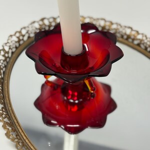 May include: A red glass candle holder with a flower-like design, holding a white candle. The candle holder sits on a mirrored surface within a gold-colored, ornate, circular frame. The candle holder's design is reflected in the mirror.