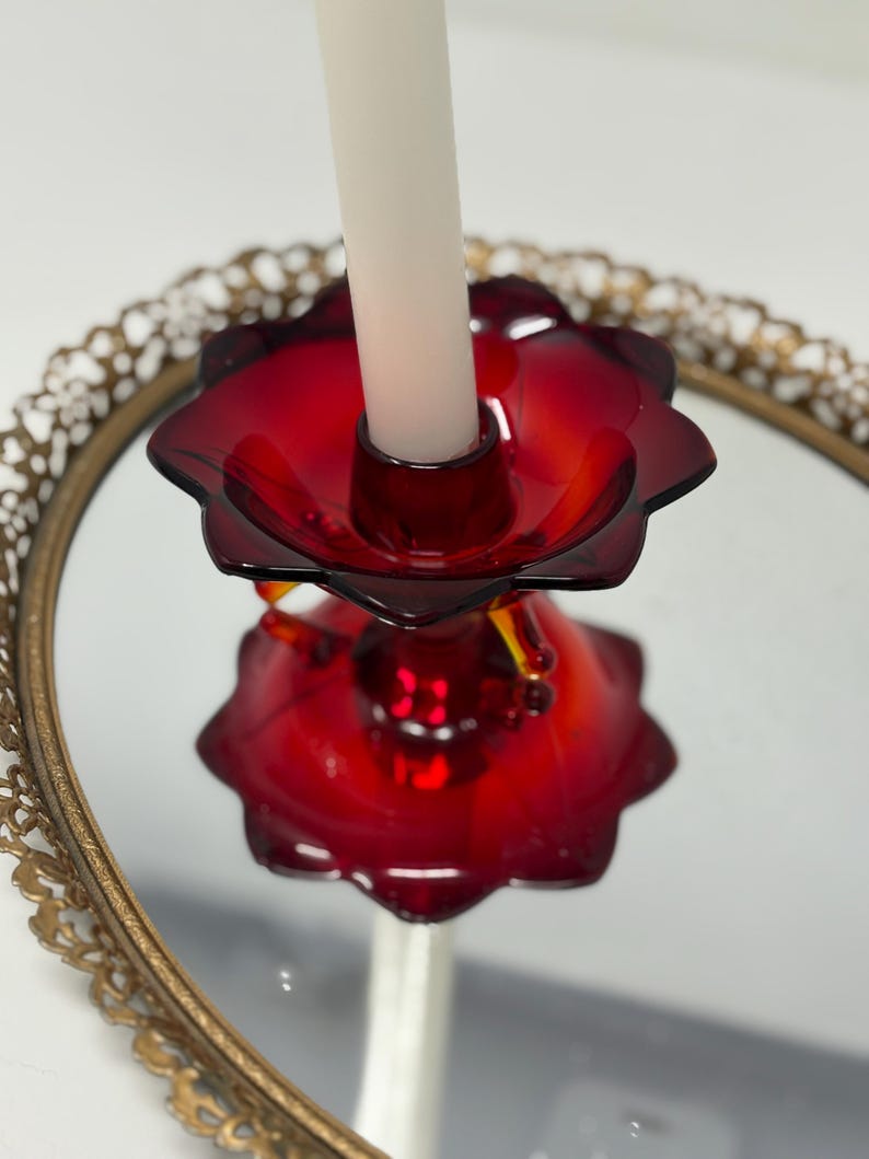 May include: A red glass candle holder with a flower-like design, holding a white candle. The candle holder sits on a mirrored surface within a gold-colored, ornate, circular frame. The candle holder's design is reflected in the mirror.