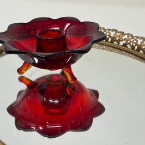 May include: A ruby red glass candle holder with a flower-shaped bowl and a base. The candle holder has three small, curved legs and a small, round candle cup. The glass has a glossy finish and is set on a decorative gold-toned tray.