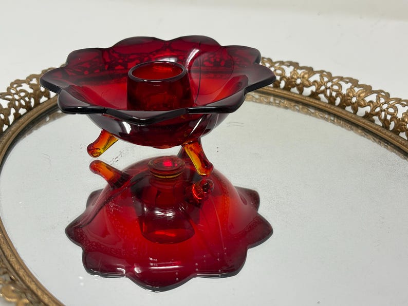 May include: A ruby red glass candle holder with a flower-shaped bowl and a base. The candle holder has three small, curved legs and a small, round candle cup. The glass has a glossy finish and is set on a decorative gold-toned tray.