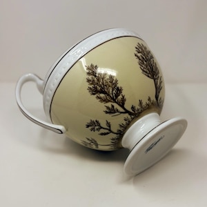 Puede incluir: Una jarra de crema de cerámica blanca con base y asa. La jarra tiene un fondo crema con un diseño de árbol marrón.