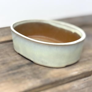 Peut inclure: Un bol ovale en céramique avec un extérieur vert clair et un intérieur marron. Le bol a une finition lisse et vernie et repose sur une surface en bois. Le bord est d'une teinte plus claire de vert.