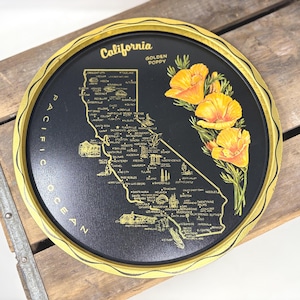 May include: A round black metal tray with a gold-coloured rim featuring a map of California. The design includes the text California and Golden Poppy, a graphic of orange poppy flowers, and a detailed map with city names and landmarks.